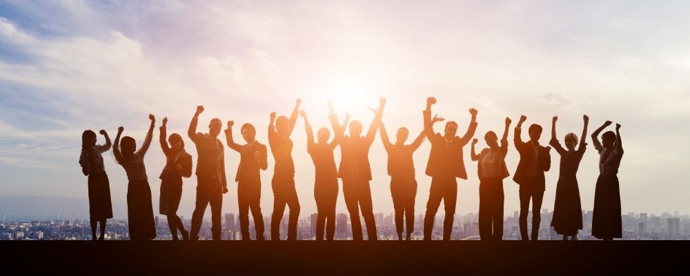 Image of a line of people standing on top of a building with the sun shining behind them. The light behind them turns the figures into silhouettes, so you cannot see their features. They all have their arms raised as if they are cheering.