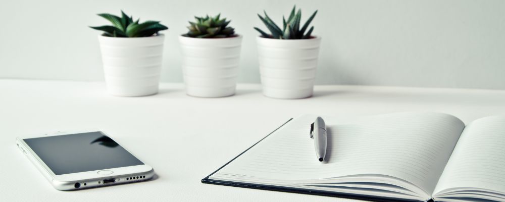 Image of a journal on a white table, with an iphone lying next to it, there are three white plant pots with succulents against a white wall. This is image is about journaling your yearly themes.